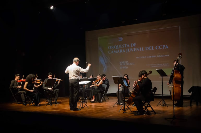 Músicos en ropa formal negra interpretan en concierto, con el director de espaldas levantando la batuta y un proyector al fondo.