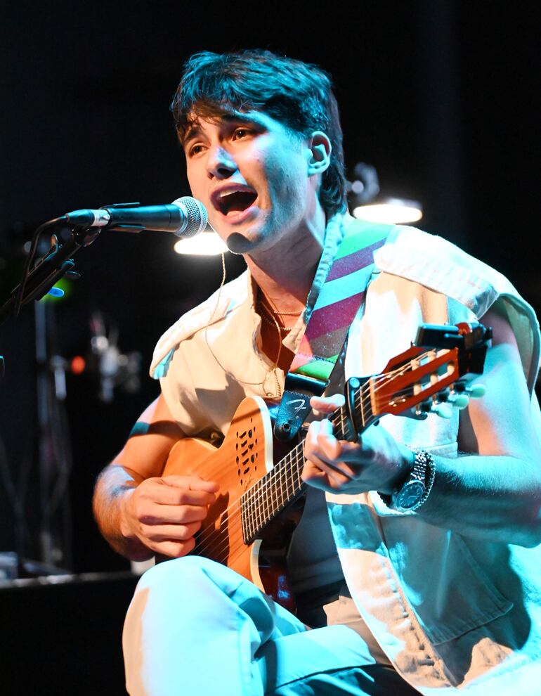 Juan Ignacio "Ancho Laterza" con guitarra acústica, sentado en escenario iluminado con luces azules, luciendo vestimenta ligera y pulseras.