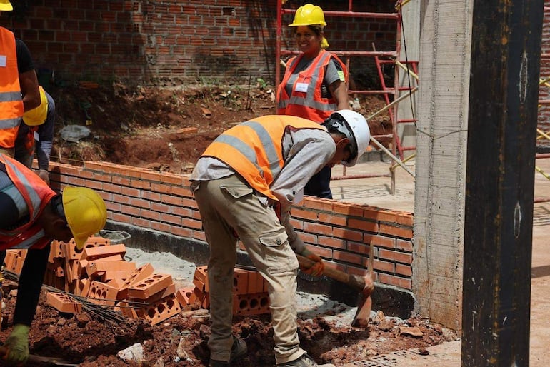 Cinco trabajadores, con cascos y chalecos naranjas, construyen una pared con ladrillos, en un ambiente de trabajo exterior.