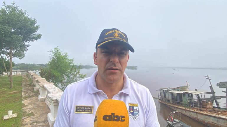 Emigdio Zayas con camiseta blanca y gorra de la Armada Paraguaya, sosteniendo un micrófono frente al río y embarcaciones amarradas.