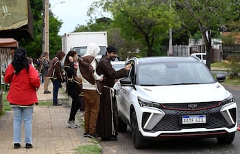 Grupo de Hermanos Capuchinos en túnicas marrones bendiciendo vehículos en una calle residencial con árboles alrededor.