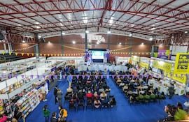 Personas sentadas en auditorio escuchan a oradores en evento decorado con tonos morados y amarillos, rodeados de stands de libros.