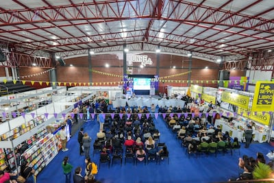 Personas sentadas en auditorio escuchan a oradores en evento decorado con tonos morados y amarillos, rodeados de stands de libros.