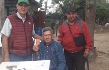 Héctor Cáceres, (Chaleco granate y quepis verde) candidato de la ANR a la Intendencia de Humaitá, posa junto a un adulto mayor y un candidato a Concejal durante la entrega de tarjetas para el cobro de la pensión destinada a personas mayores.