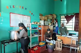 Mujer de espaldas removiendo olla, otra pelando frutas y hombre cortando verduras en cocina comunitaria con estantes verdes.