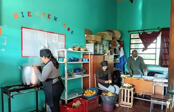 Mujer de espaldas removiendo olla, otra pelando frutas y hombre cortando verduras en cocina comunitaria con estantes verdes.