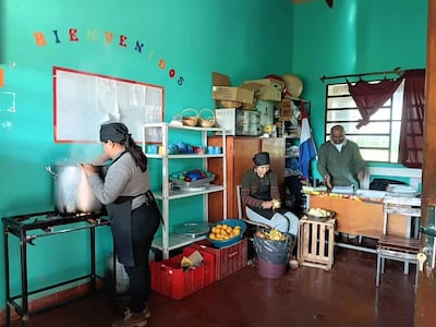Mujer de espaldas removiendo olla, otra pelando frutas y hombre cortando verduras en cocina comunitaria con estantes verdes.