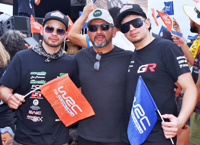 Tres hombres con gafas de sol y camisetas oscuras sostienen banderas del WRC en un evento festivo al aire libre.