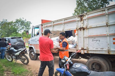 Dos hombres en camiseta roja y chaleco naranja interactúan mientras reciben paquetes de víveres al aire libre.