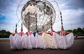 Siete mujeres sonrientes en camisetas de rayas rojas y blancas y faldas blancas, posan junto al Unisphere en un ambiente festivo.