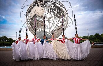 Siete mujeres sonrientes en camisetas de rayas rojas y blancas y faldas blancas, posan junto al Unisphere en un ambiente festivo.