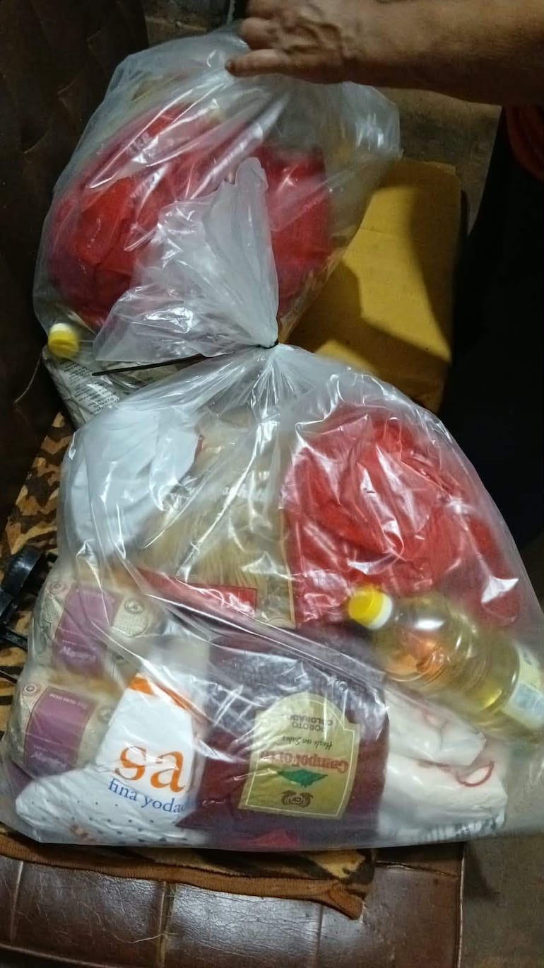 Mano sostiene una bolsa de víveres, rodeada de productos alimenticios en un ambiente interior con sofá marrón.