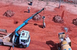 Trabajadores en overoles y cascos, uno levantando un recipiente con grúa, en terreno rojo de construcción.