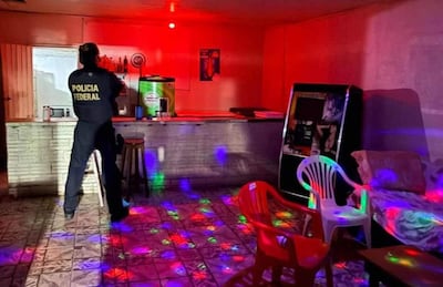 Agente de Policía Federal de espaldas, con uniforme, frente a un bar luminoso con sillas de colores y una barra en el fondo.