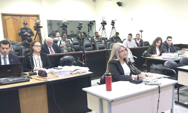 Mujer de cabello largo y rubio hablando al micrófono, rodeada de hombres y mujeres atentos en un ambiente formal de tribunal.