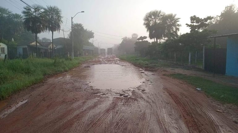 Calle rural con vegetación y casas simples, mostrando charcos de agua y neblina matutina, sin personas visibles.