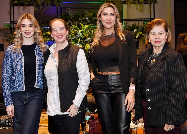 Cuatro mujeres sonrientes, una con chaqueta azul, gafas y blusa negra, otra de cabello lacio en blusa negra, y una de rayas.