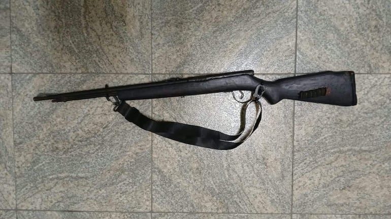Rifle negro con correa de transporte sobre fondo de losas de cerámica en un ambiente interior.