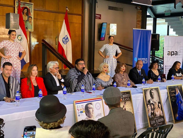 Alda Cardozo en elegante vestido habla en mesa con artistas, rodeada de público y banderas paraguayas.