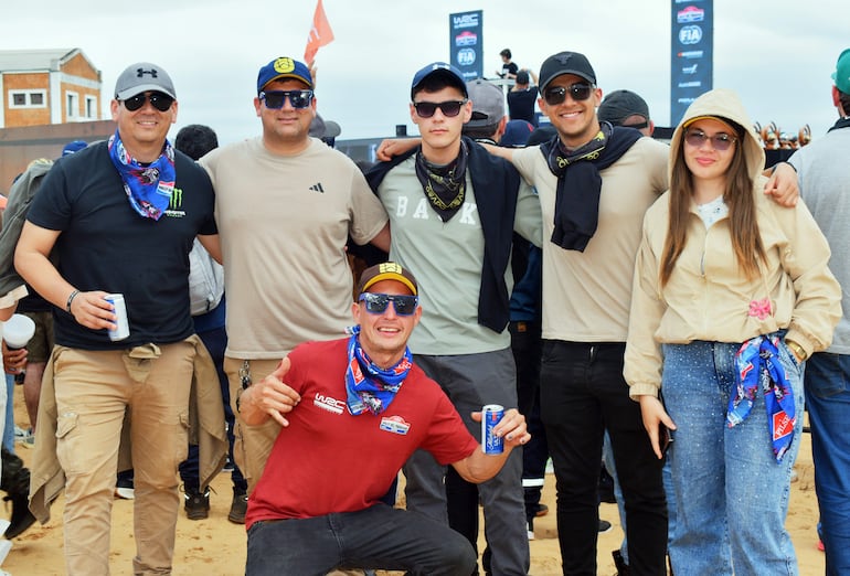 Grupo de seis personas con ropa casual sonríen en evento, uno agachado con bebida en mano y gafas de sol.