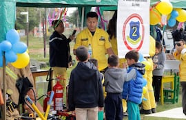 Niños atentos a un bombero en uniforme amarillo, rodeados de globos y decoraciones en la plaza. Una mujer graba con su móvil.