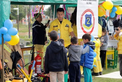 Niños atentos a un bombero en uniforme amarillo, rodeados de globos y decoraciones en la plaza. Una mujer graba con su móvil.
