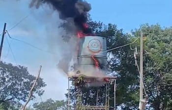 Tanque de agua en llamas con densa columna de humo negro en un entorno de vegetación y postes de electricidad.