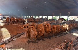 Ambiente festivo con personas de pie al fondo, mientras se cocinan costillas en estructuras metálicas rodeadas de leña humeante.