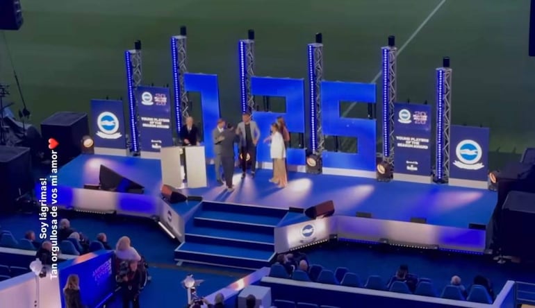 Diego Gómez, vestido formal, recibe un premio en el centro de un escenario iluminado rodeado de presentadores y una audiencia.