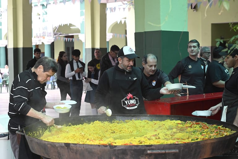 Hombre mayor con cabello canoso y camisa de rayas sirve comida, mientras otros hombres jóvenes colaboran en un ambiente festivo.