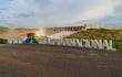 Cartel de 'ITAIPU NACIONAL' en primer plano, con represa y paisaje natural al fondo bajo un cielo nublado.