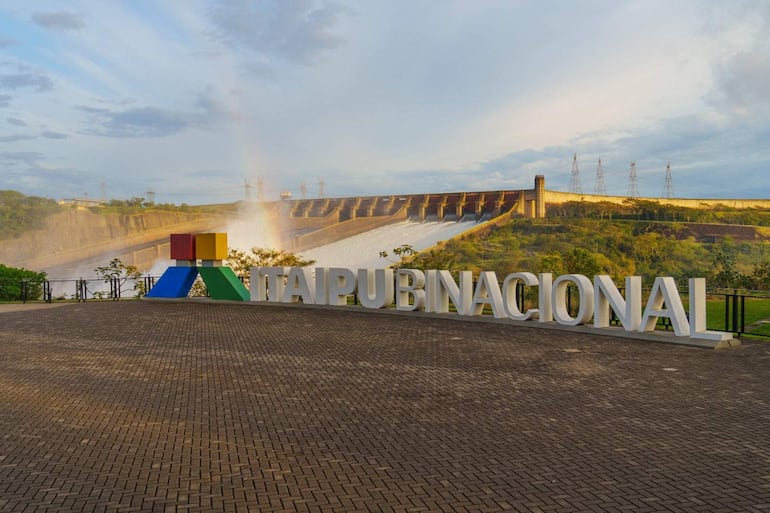Cartel de 'ITAIPU NACIONAL' en primer plano, con represa y paisaje natural al fondo bajo un cielo nublado.