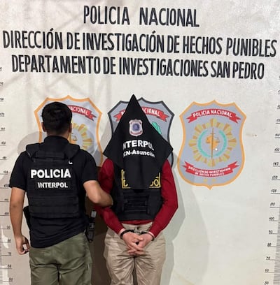 Agente de policía interactuando con David Wiebe Martens, quien lleva una capucha negra y camiseta roja, en comisaría.