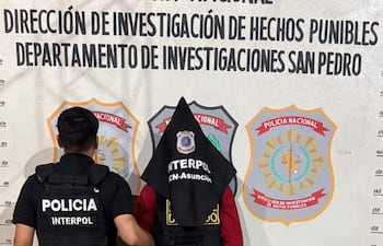 Agente de policía interactuando con David Wiebe Martens, quien lleva una capucha negra y camiseta roja, en comisaría.