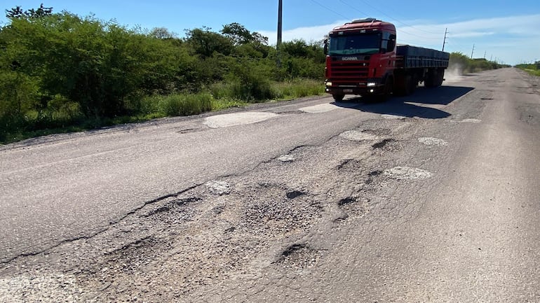 Camión rojo Scania levantando polvo en una carretera deteriorada, con vegetación baja a los lados y cielo despejado.