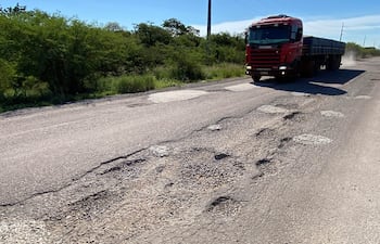 Camión rojo Scania levantando polvo en una carretera deteriorada, con vegetación baja a los lados y cielo despejado.