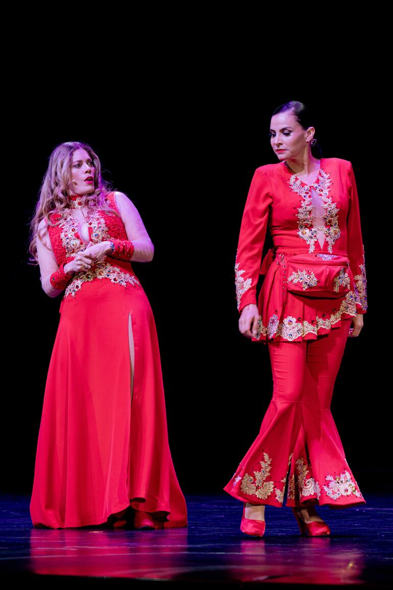 Lorna Cepeda con cabello rubio y vestido rojo, y Natalia Ramírez con cabello recogido y vestido rojo, en un escenario teatral oscuro.