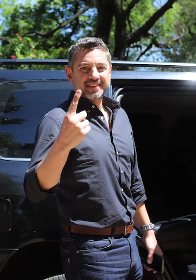 Pedro Alliana, con cabello canoso y camisa negra, levanta un dedo señalando, frente a un vehículo oscuro en un entorno urbano.