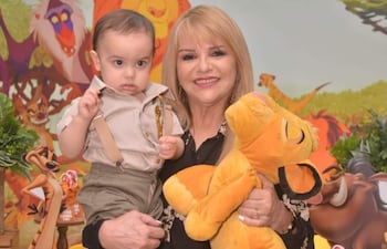 Mujer rubia con vestido oscuro sonríe mientras sostiene a un niño serio que señala, rodeados de decoraciones de 'El Rey León'.