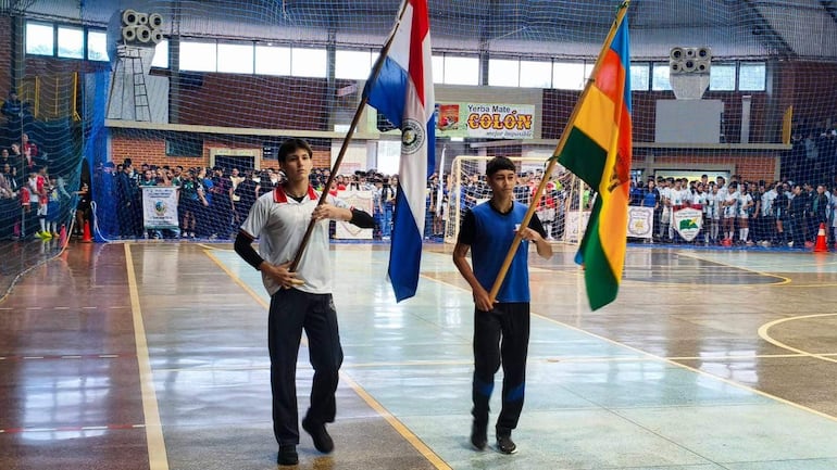 Dos jóvenes marchan con banderas de Paraguay y Bolivia, rodeados de un público animado en un gimnasio.