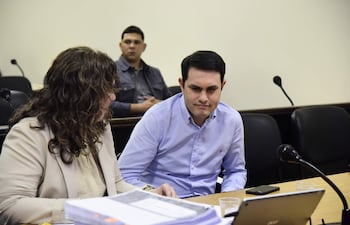 Hernán Rivas habla con cabello rizado, mientras un hombre de camisa de cuadros observa pensativo en un tribunal.
