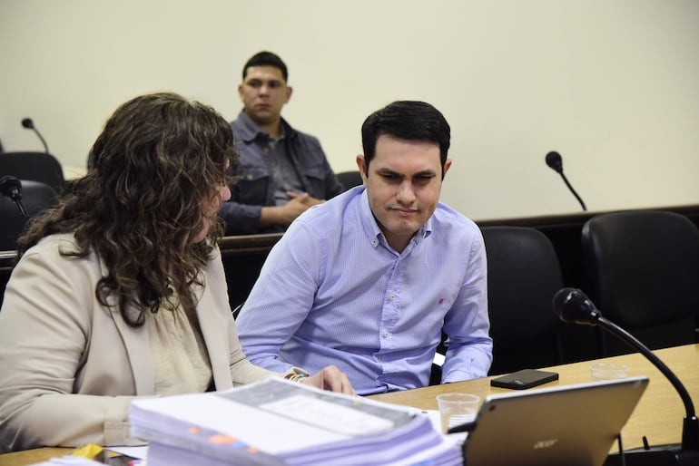 Hernán Rivas habla con cabello rizado, mientras un hombre de camisa de cuadros observa pensativo en un tribunal.
