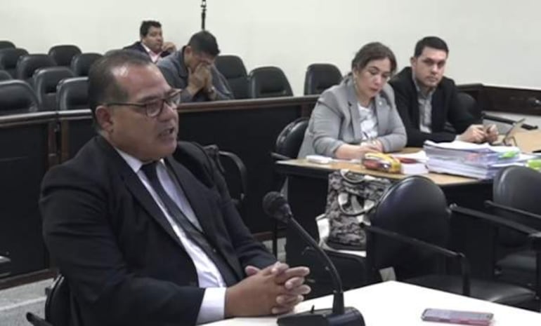 Hombre con traje oscuro y gafas declaran en audiencia; tres personas escuchan, una con chaqueta gris y otra evaluando documentos.