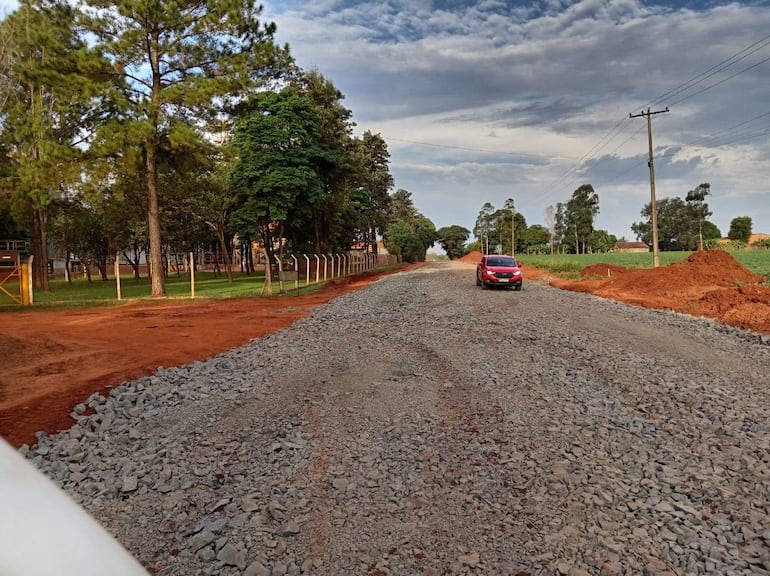 Carretera en construcción con grava y tierra, un coche rojo avanza por el camino rodeado de árboles altos.