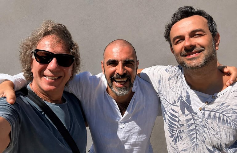 Tres hombres sonrientes: uno con gafas de sol y camiseta azul, otro con camisa blanca y el tercero en camiseta clara.