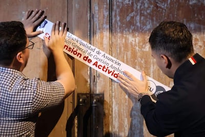 Dos hombres de perfil fijan un cartel que dice 'Municipalidad de Asunción' y 'Suspensión de Actividad' en una puerta de madera.