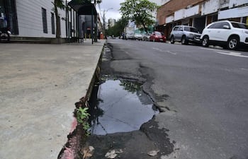 Calle inundada con agua reflejando vehículos estacionados y una acera desgastada en un día nublado.