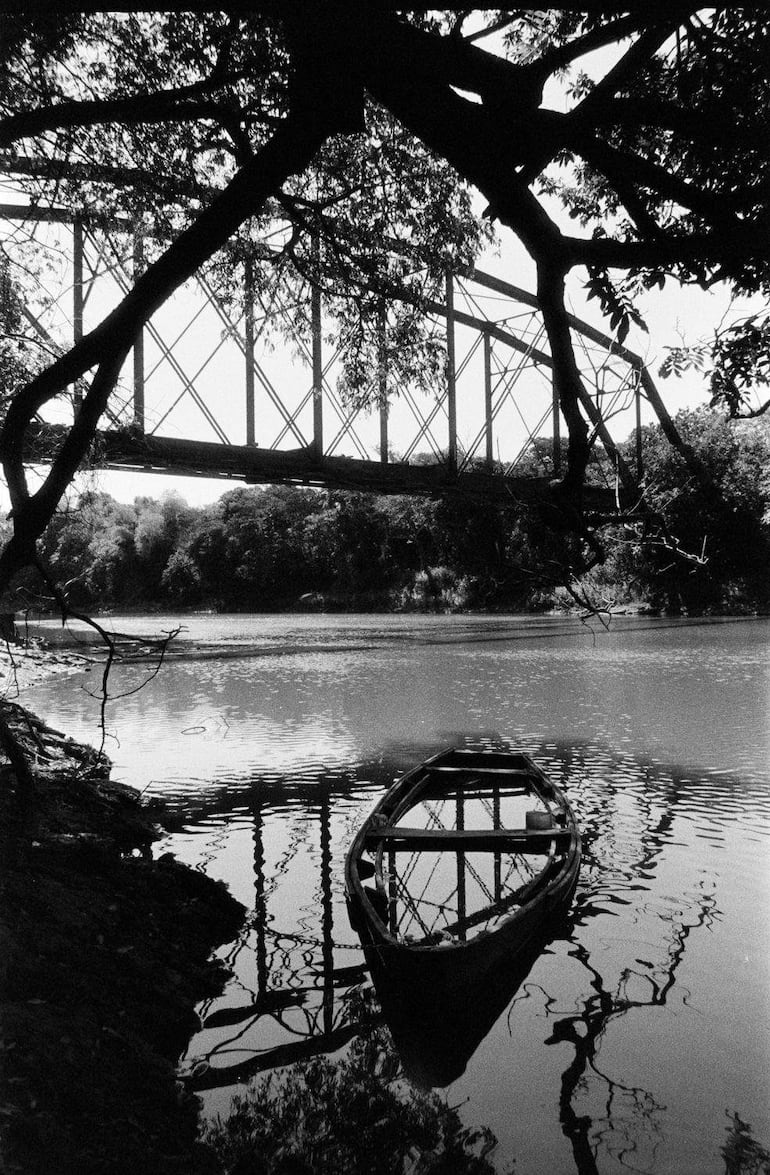 Paisaje en blanco y negro con un puente metálico y un bote de remos anclado en la orilla, rodeado de vegetación densa.