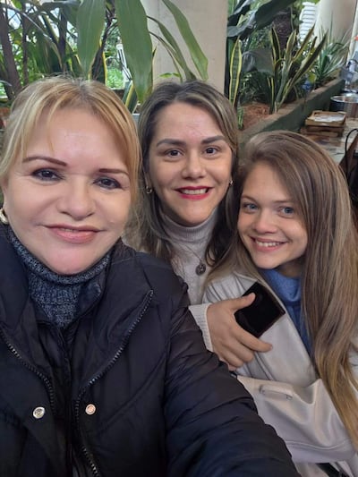 Tres mujeres sonrientes en un café: una con abrigo negro, otra en blusa blanca y suéter gris, y otra con chaqueta clara.