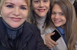 Tres mujeres sonrientes en un café: una con abrigo negro, otra en blusa blanca y suéter gris, y otra con chaqueta clara.